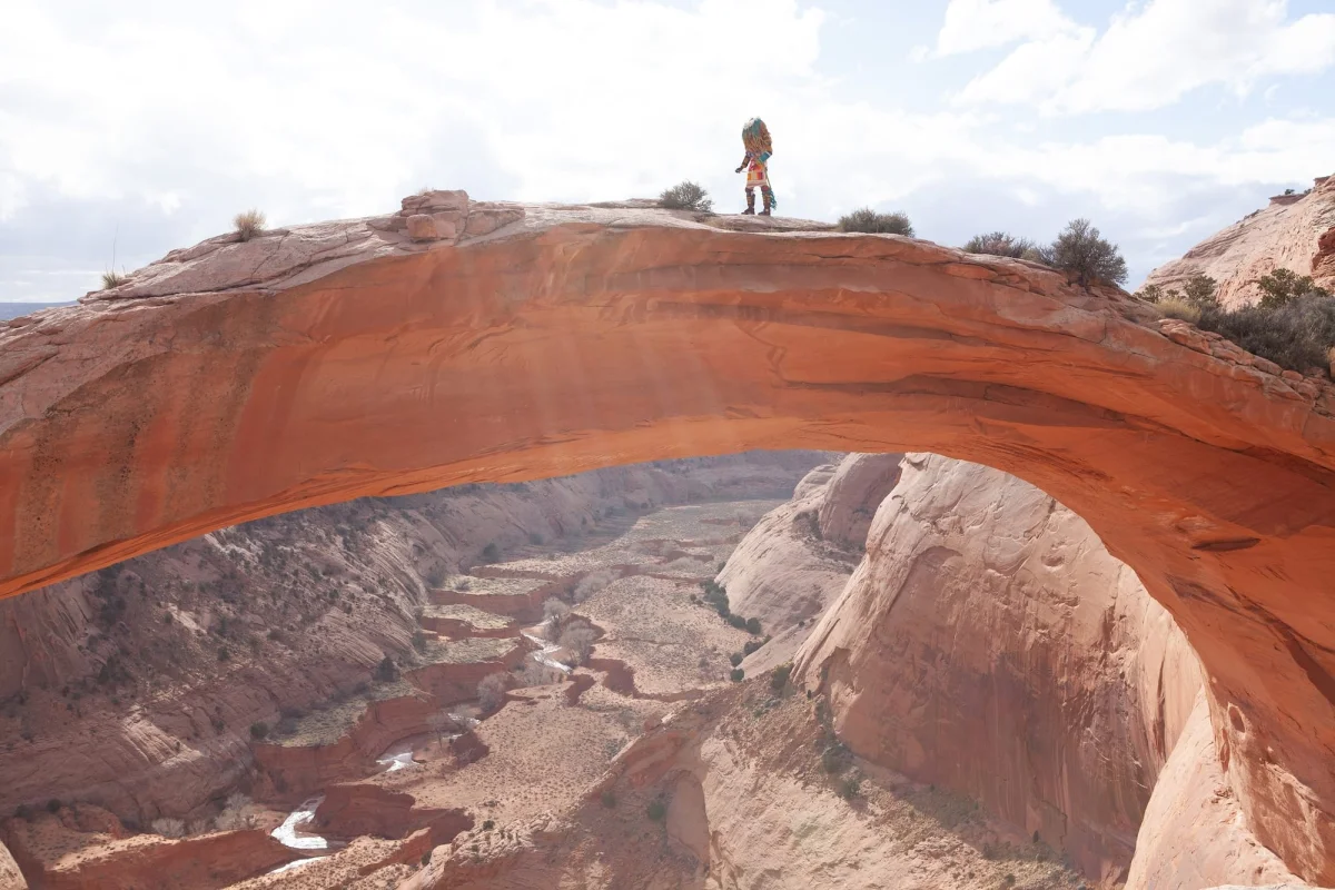 Cannupa Hanska Luger wears a vibrant costume informed by Indigenous narratives and walks across a natural sandstone arch on the Navajo Nation