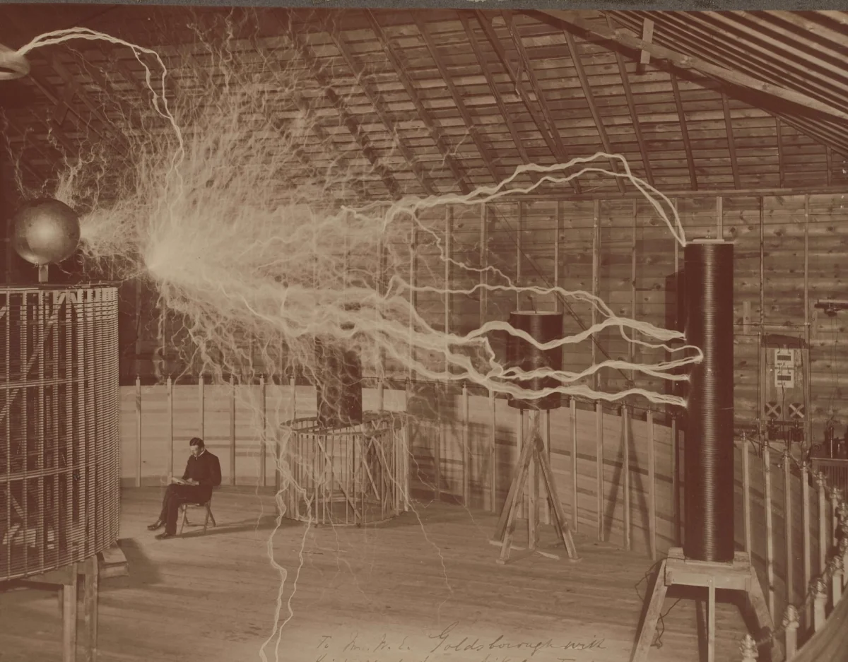 a double-exposure of Nikola Tesla seated in a laboratory while enormous electrical currents blast through the space