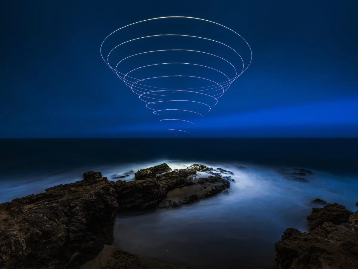 a "light painting" photograph with a cone of swirling light, created using a drone, over a dramatically lit nighttime coastal landscape