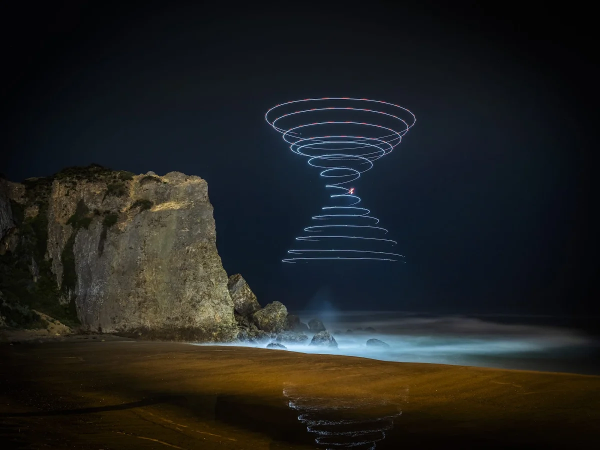 a "light painting" photograph with a double cone of swirling light, created using a drone, over a dramatically lit nighttime desert landscape with a natural pool that glows under the light